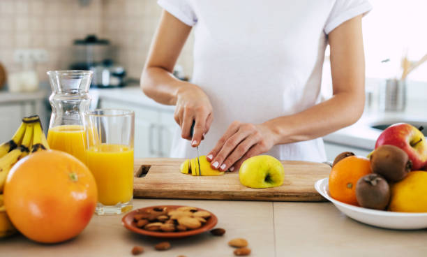 cute beautiful and happy young brunette woman in the kitchen at home is preparing fruit vegan salad or a healthy smoothie and having fun - routine foods and drinks clothing household equipment imagens e fotografias de stock