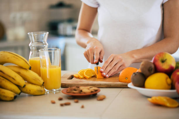 cute beautiful and happy young brunette woman in the kitchen at home is preparing fruit vegan salad or a healthy smoothie and having fun - routine foods and drinks clothing household equipment imagens e fotografias de stock