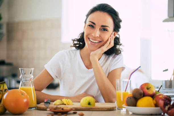 cute beautiful and happy young brunette woman in the kitchen at home is preparing fruit vegan salad or a healthy smoothie and having fun - routine foods and drinks clothing household equipment imagens e fotografias de stock
