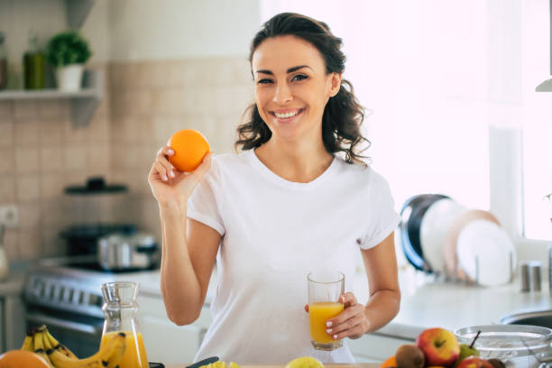 cute beautiful and happy young brunette woman in the kitchen at home is preparing fruit vegan salad or a healthy smoothie and having fun - routine foods and drinks clothing household equipment imagens e fotografias de stock