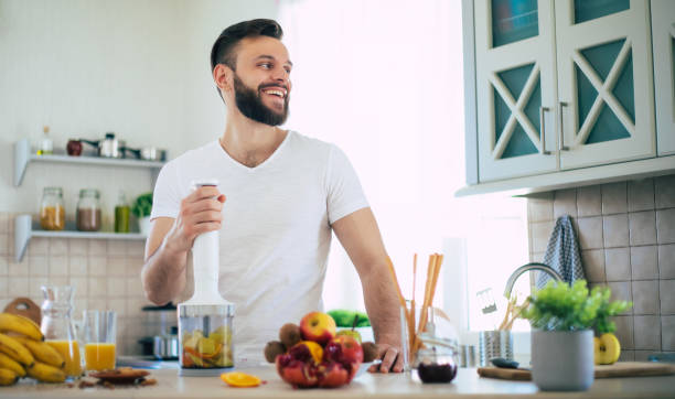 przystojny młody sportowy uśmiechnięty mężczyzna w kuchni przygotowuje wegańską zdrową sałatkę z owoców i smoothie w dobrym nastroju - blender salad lemon vegetable zdjęcia i obrazy z banku zdjęć