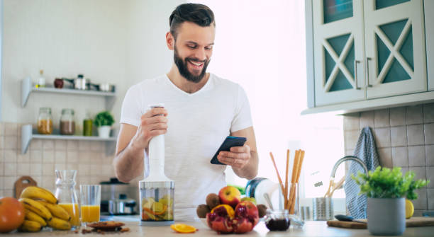 handsome young sporty smiling man in the kitchen is preparing vegan healthy fruits salad and smoothie in a good mood - blender apple banana color image imagens e fotografias de stock