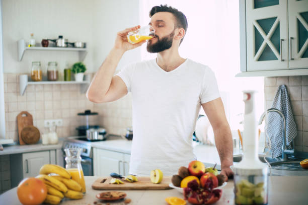 il bel giovane uomo sportivo sorridente in cucina sta preparando insalata di frutta sana vegana e frullato di buon umore - juice glass healthy eating healthy lifestyle foto e immagini stock