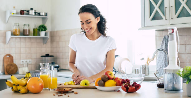 cute beautiful and happy young brunette woman in the kitchen at home is preparing fruit vegan salad or a healthy smoothie and having fun - routine foods and drinks clothing household equipment imagens e fotografias de stock