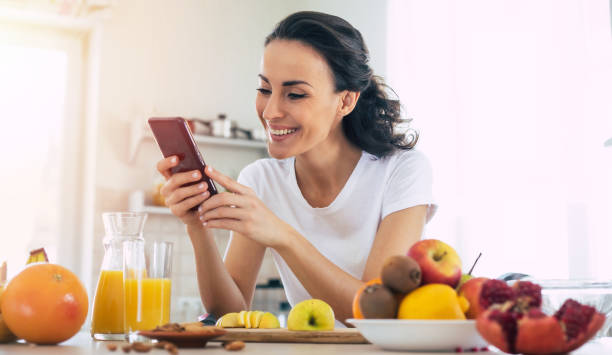 la femme mignonne de sourire utilise le smartphone dans la cuisine à la maison tout en cuisinant la salade végétalienne de fruits - healthy eating breakfast ripe fruit photos et images de collection