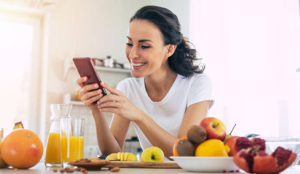 uśmiechnięta urocza kobieta używa smartfona w kuchni w domu podczas gotowania owoców wegańskiej sałatki - routine foods and drinks clothing household equipment zdjęcia i obrazy z banku zdjęć