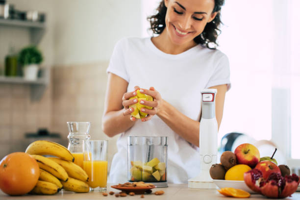 cute beautiful and happy young brunette woman in the kitchen at home is preparing fruit vegan salad or a healthy smoothie and having fun - routine foods and drinks clothing household equipment imagens e fotografias de stock