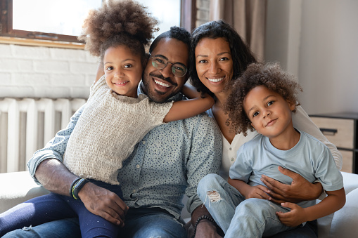 Happy friendly family. Portrait of smiling pleasant black married couple sitting on sofa at living room embracing warm looking at camera holding two little kids elder daughter and younger son on laps