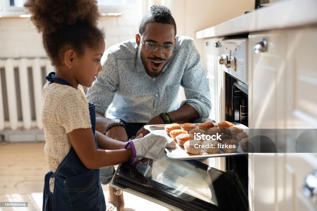 Excited african dad watching daughter taking muffins out of oven Wow, looks great! Excited caring millennial african dad or grown elder brother watching concentrated small black daughter or younger preteen sister taking pan with self baked tasty muffins out of oven Family Stock Photo