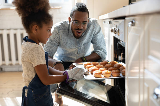 Excited african dad watching daughter taking muffins out of oven Wow, looks great! Excited caring millennial african dad or grown elder brother watching concentrated small black daughter or younger preteen sister taking pan with self baked tasty muffins out of oven family home stock pictures, royalty-free photos & images