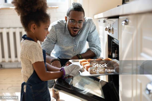 Excited African Dad Watching Daughter Taking Muffins Out Of Oven Stock Photo - Download Image Now