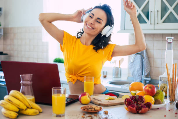 excited beautiful happy young woman in headphones is dancing and cooking in the kitchen at home - routine foods and drinks clothing household equipment imagens e fotografias de stock