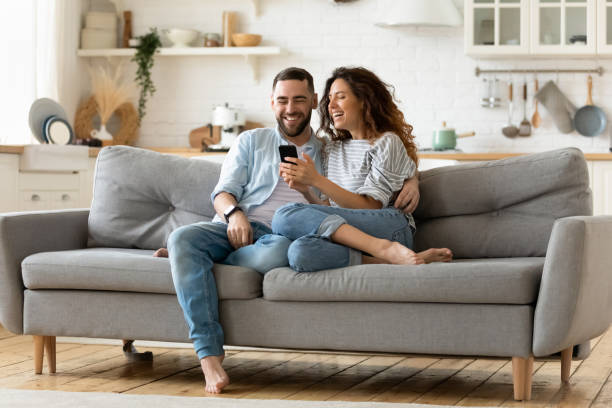 Happy young woman and man hugging, using smartphone together Happy young woman and man hugging, using smartphone together, sitting on cozy couch at home, smiling overjoyed wife and husband looking at phone screen, sitting on sofa in modern living room couple stock pictures, royalty-free photos & images