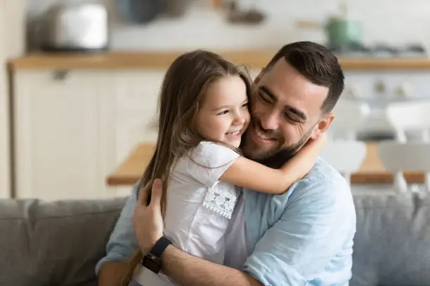 Close up smiling loving young father hugging adorable little daughter, enjoying tender moment, spending weekend together, sitting on cozy couch at home, good family relationship between dad and child
