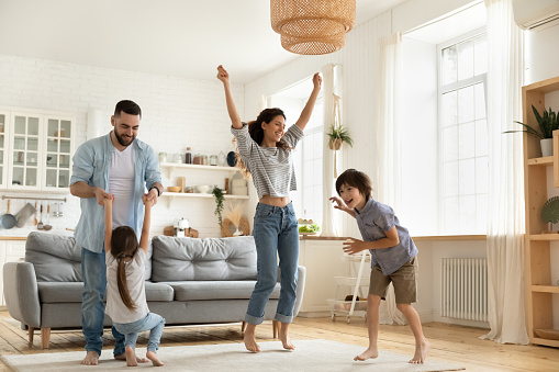 Happy parents with kids dancing in modern living room, having fun, playing active game, overjoyed mother and father with children siblings dancing to favorite music, jumping, celebrating event