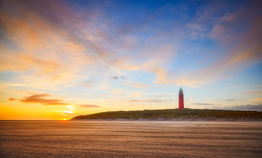 The lighthouse of Texel during sunset seen from the beach. The lighthouse is located in the dunes. The sky is clear with some light clouds in it. Only a part of the sun can still be seen just above the horizon.