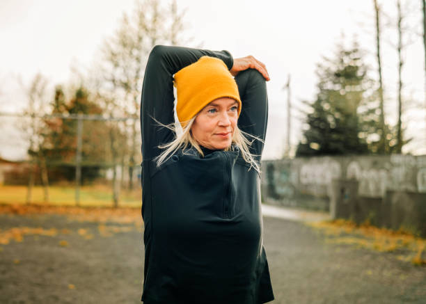 Jogger doing arm stretches in morning Mature woman stretching arms in the city park. Female jogger wearing knitted hat doing warm up workout outdoor on a winter morning. active lifestyle stock pictures, royalty-free photos & images