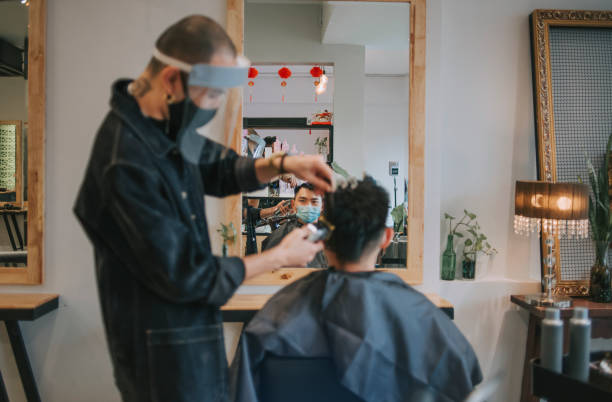 riflessione parrucchiere cinese asiatico con abbigliamento da lavoro protettivo che taglia i capelli per il suo cliente nel salone dei capelli - shaving men electric razor reflection foto e immagini stock