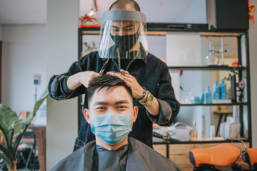 Asian chinese hairstylist with protective workwear cutting hair for his customer in hair saloon