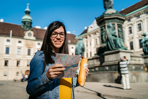 Young Caucasian beautiful tourist woman exploring Vienna with map. Eating wiener würstchen / sausage hot dog.