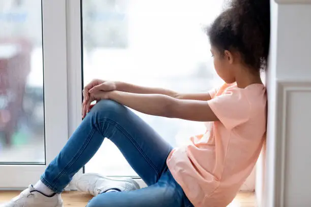 Photo of Sad african teenage girl sitting on window sill at home