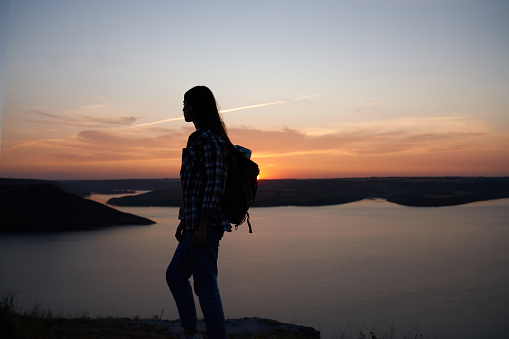Silhouette of young woman in casual clothing with backpack on shoulder standing on mountain peak. Background of beautiful sunset over Bakota bay.