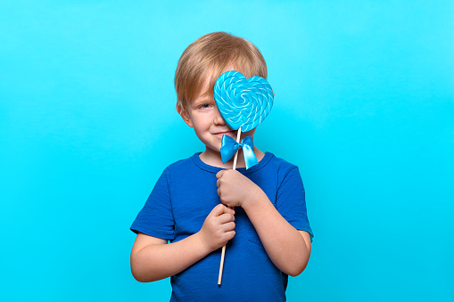 Caucasian child hold huge heart shaped lollipop on stick, close his eyes and smile. Studio shot with copy space on blue solid color background, isolated. Concept of sweets, nutrition and childhood.