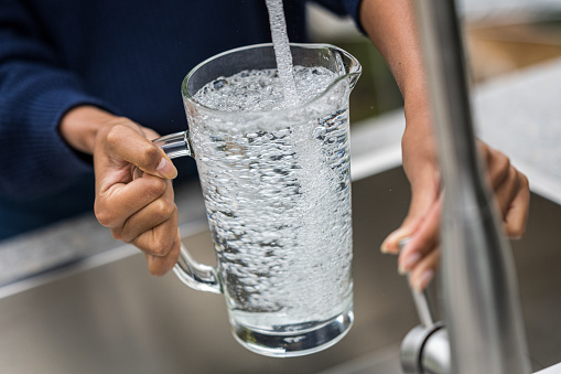Close-up of female hands holding big glass jar and filling it in with water from faucet. Water usage concept.