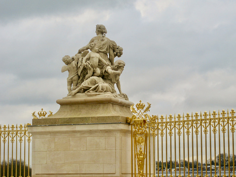 Horizontal closeup of the exterior gold-painted fence and the rear view of a sculpture mounted on a plinth in the grounds of the Palace of Versailles