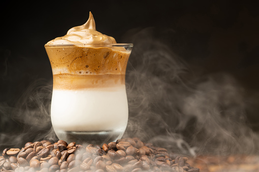 Ice Dalgona Coffee, Korean Whipped Drink in a Transparent Glass, Stands on Coffee Beans on a Dark Background with Smoke. Close-up, Copy Space