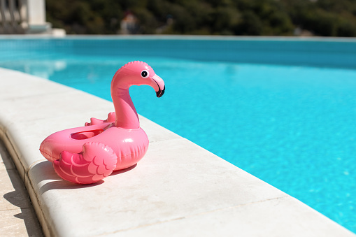 Inflatable pink mini flamingo, cocktail stand near swimming pool on bright sunny day, copy space. Concept of summer vacation, entertainment, water, air, sunbathing, health. Side view. Horizontal.