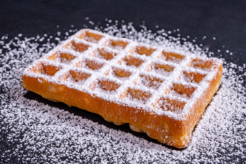 Sweet waffles sprinkled with powdered sugar prepared for serving. A tasty dessert eaten with fruit and preserves. Dark background.