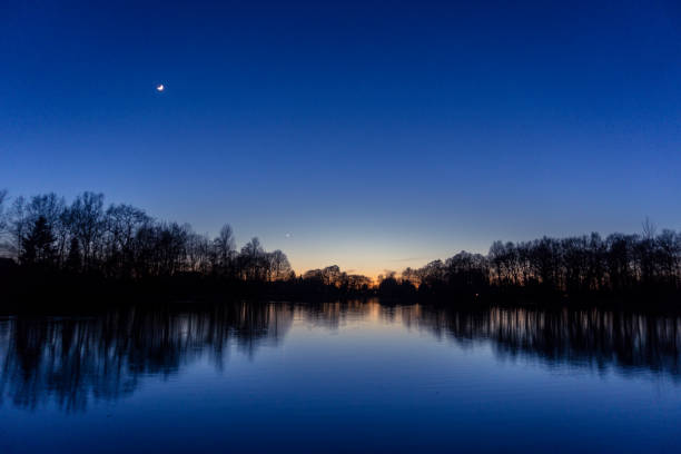 Clear blue sky after sunset at lake with reflecting silhouettes of trees, venus and crescent moon Clear blue sky after sunset at lake with reflecting silhouettes of trees, venus and crescent moon blue hour twilight photos stock pictures, royalty-free photos & images