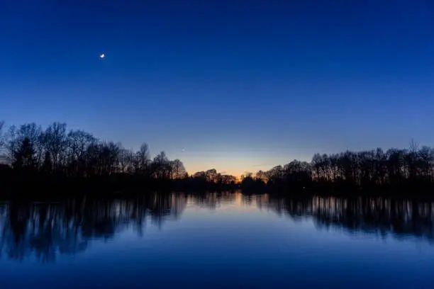 Clear blue sky after sunset at lake with reflecting silhouettes of trees, venus and crescent moon