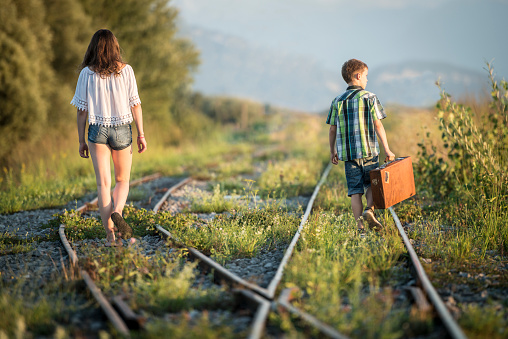 Rear view of a teenage girl and a boy holding a suitcase. They are walking on different rails behind a switch.