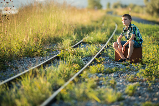 Front view of a boy sitting on a suitcase waiting for the train beside the tracks in the  evening sun.