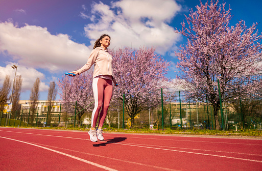 Female athlete exercising outdoors with a jumping rope. She is doing a sports training on running track. Sunny early spring day.