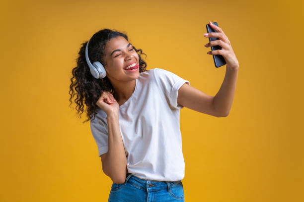 音楽を聴いて、彼女の携帯電話で自分撮りを取るパーフェクサー笑顔を持つ魅力的な陽気な暗い肌の女の子 - skin singer ストックフォトと画像