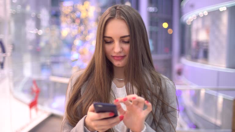 Young woman surfs internet or texting sms message using modern smartphone over blurry big mall background. 4 k