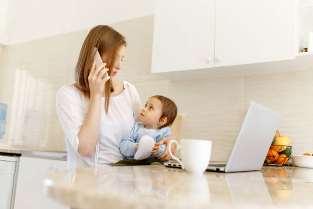 istock Beautiful business mom is talking on the mobile phone and taking notes while spending time with her cute baby boy at kitchen home office. Working mother concept,Laptop on table 1309167251