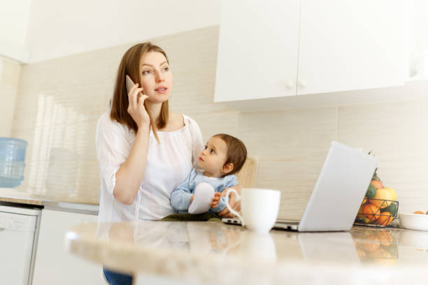 istock Beautiful business mom is talking on the mobile phone and taking notes while spending time with her cute baby boy at kitchen home office. Working mother concept,Laptop on table 1309167261