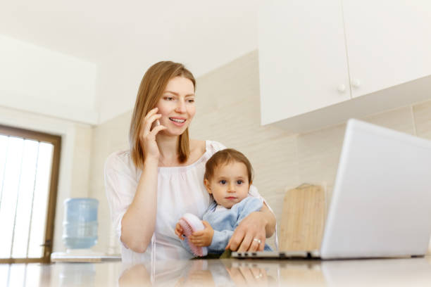 istock Beautiful business mom is talking on the mobile phone and taking notes while spending time with her cute baby boy at kitchen home office. Working mother concept,Laptop on table 1309167266
