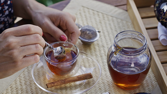 Close-up view of young woman hands pouring honey natural sweetener into hot tea cup on a table at outdoors terrace