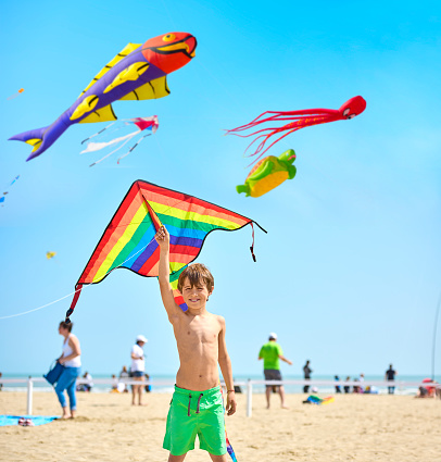 Child with swimsuit plays on the beach by the sea with a colorful kite. In the background, many people are walking on the beach.