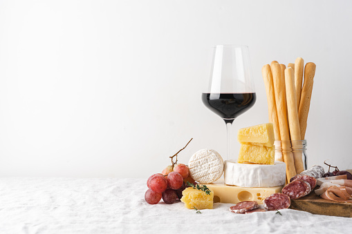 Glass of red wine and cheese, sausage, grapes and grissini bread sticks on the table. Light background, traditional wine snacks on tablecloth covered table