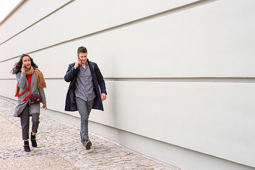 A woman and a man on the phone while walking on a sidewalk alongside a building