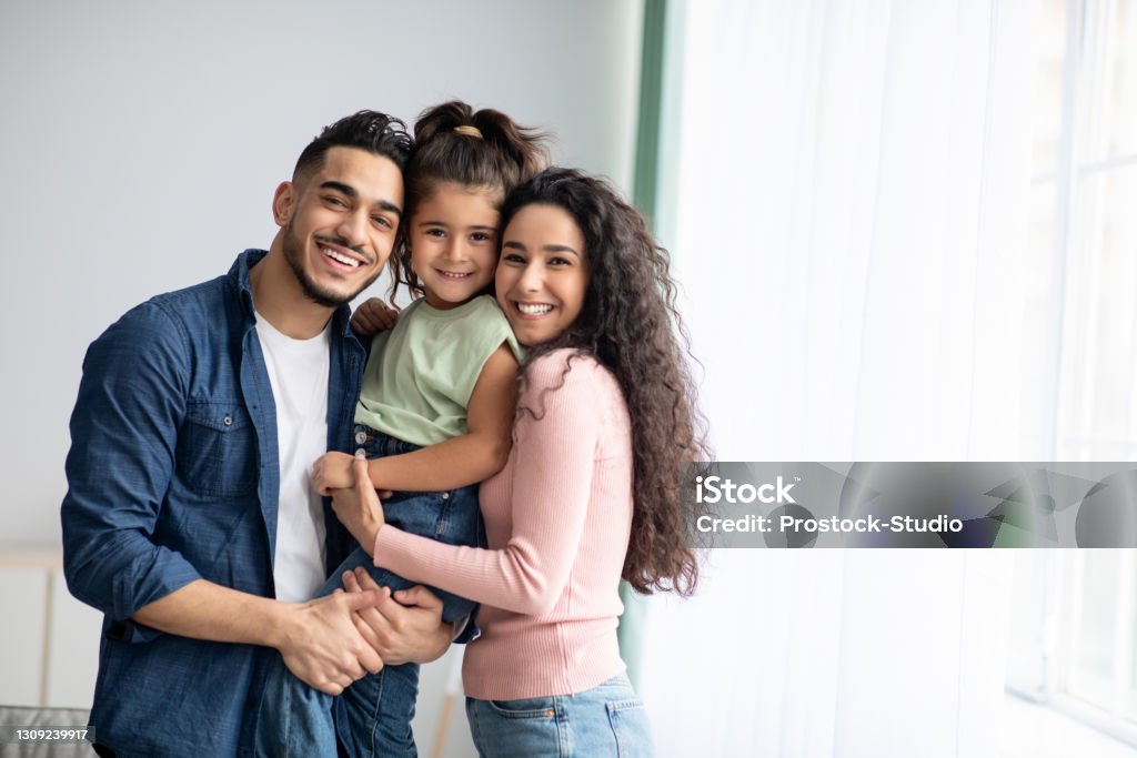 Portraif Of Happy Arabic Parents Posing With Their Little Daughter At Home Portraif Of Happy Arabic Parents Posing With Their Little Daughter At Home, Loving Middle Eastern Family Of Three Embracing Near Window And Smiling At Camera, Enjoying Time Together, Copy Space Family Stock Photo