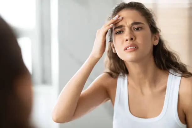 First Wrinkle. Concerned Woman Searching Wrinkles Looking At Forehead And Touching Face Standing Near Mirror In Modern Bathroom Indoor. Facial Skin Problems And Aging Concept. Selective Focus