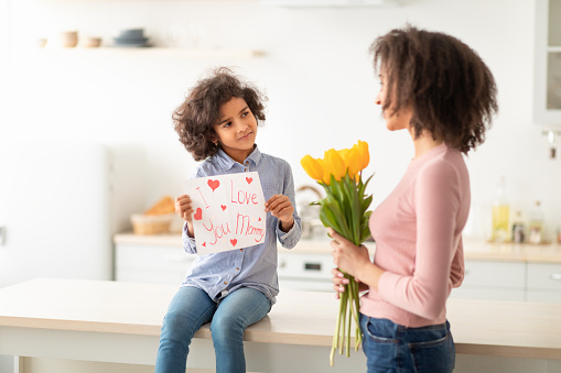 Portrait of little African American girl greeting her mum, showing I Love You, Mommy paper poster, sitting on dinner table. Happy woman holding bouquet of fresh yellow tulips flowers