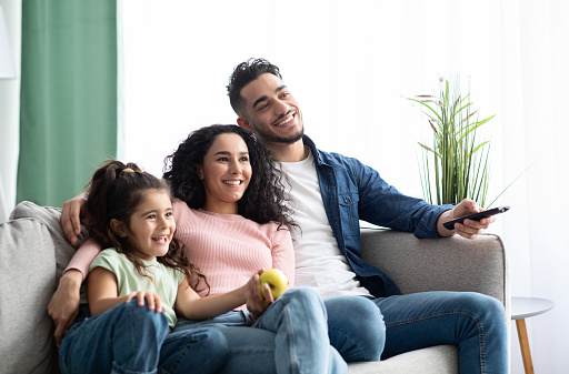 Happy arabic family of three relaxing and watching tv at home, middle eastern father, mother and little daughter resting on couch in living room, enjoying spending weekend together, copy space
